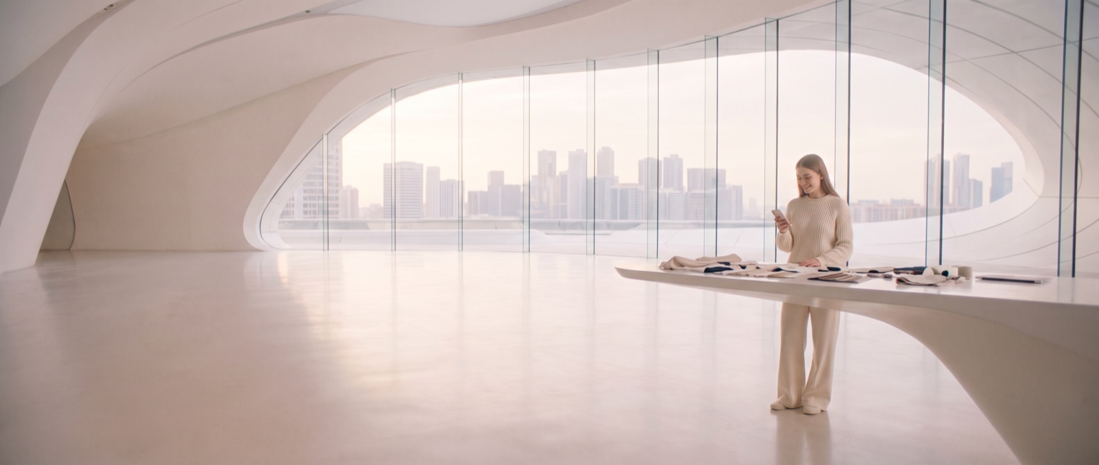 Morning in a vast white studio of flowing curves and glass above the city; a young designer smiles at her phone.